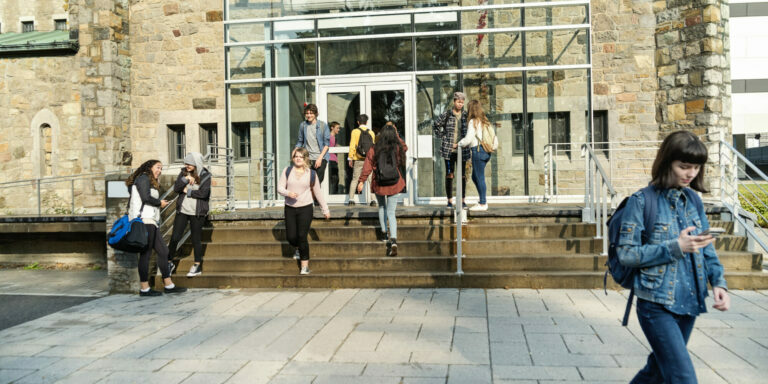 Medium group of multi-ethnic students going in and out of University-College entrance. Some are sitting and standing on a small staircase on a sunny day. Horizontal full length shot with copy space.