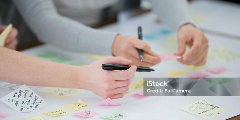 A multi-ethnic group of business professionals are working on a brainstorming board. They are using post it notes on a white board with new ideas.